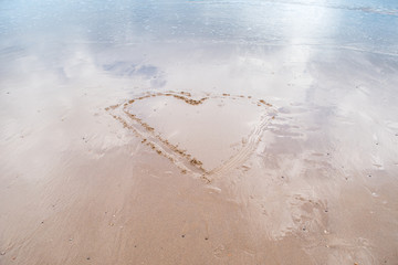 minimalist image of a large heart drawn in the sand on a flat beach with gentle tide going out and blue sky reflecting in remaining water