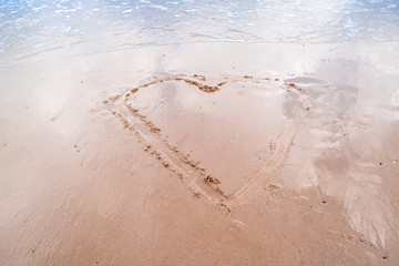 minimalist image of a large heart drawn in the sand on a flat beach with gentle tide going out and blue sky reflecting in remaining water