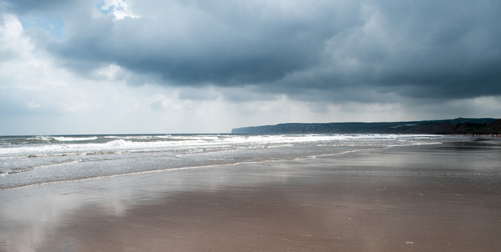 panoramic image of dark storm clouds above a flat sandy beach with white waves at Bempton Cliffs, UK in the background, depicting low colour palette, sun shining onto the sea from behind