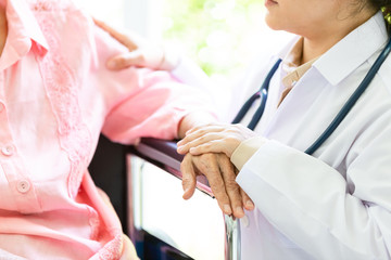 Caring doctor or nurse supporting disabled senior asian woman on wheelchair in hospital,female caregiver holding elderly patient&rsquo;s hand comforting during illness,help,health care concept