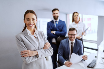 Fototapeta premium Gorgeous smiling businesswoman in suit standing in office with hands crossed. In background her successful team posing.
