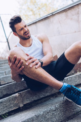 young sportsman sitting on stairs and suffering from pain while touching injured leg