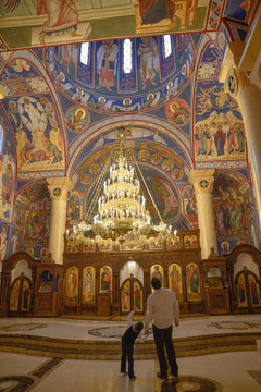 Father and son standing side by side in front of the icons of the great hall in orthodox church while the kid is pointing up and showing his dad the icons painted on the wall