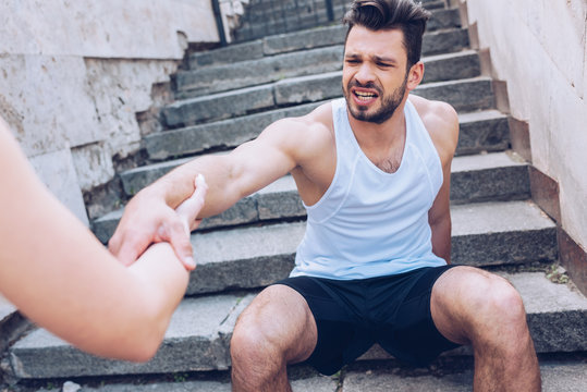 partial view of woman giving hand to injured sportsman suffering from pain while sitting on stairs