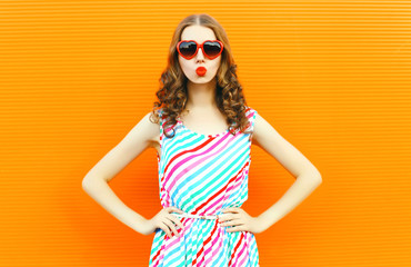 Portrait pretty woman blowing red lips sends sweet air kiss wearing red heart shaped sunglasses, colorful striped dress on orange wall background