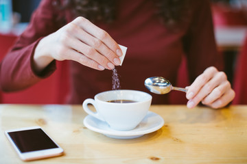 Woman's hands pouring sugar in black coffee - girl sitting at the table with espresso and smartphone - blood and glycemic index control for diabetes -excess of white sugar in food concept
