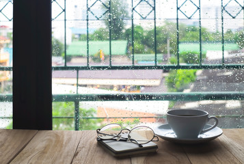 White cup of coffee with steam over a coffee cup and glasses, White cup of caffe americano on wooden table near windows in rainy morning day