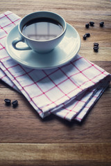 White cup of coffee with coffee beans and napkin on wooden table.