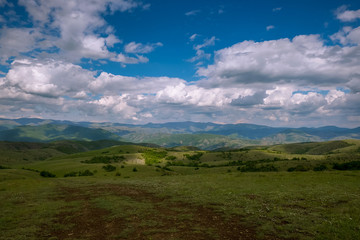 A wide green mountain field and a beautiful sunny day. In the sky are thick white robes and blue sky among them. In the distance, the surrounding mountains are visible.