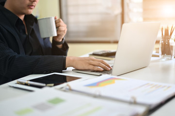 Cropped shot businessman using laptop and holding coffee mug on workplace.