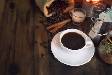 a cup of coffee americano placed on wooden table, There are stove, roasted coffee beans sag and Italian coffee maker(mocha pot)