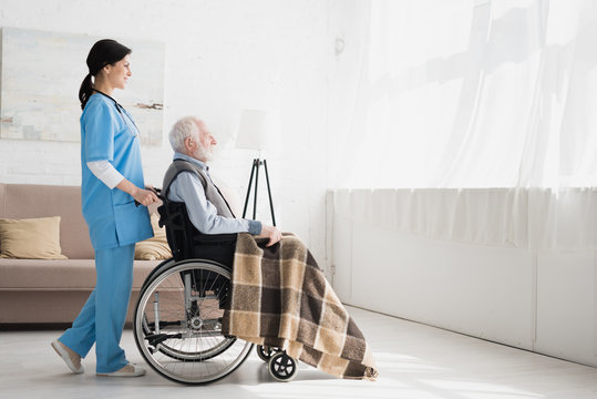 Side View Of Happy Nurse Standing In Room With Copy Space, And Carrying About Disabled Grey Haired Man In Wheelchair