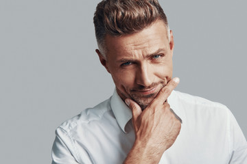 What decision to make? Handsome young man looking at camera and keeping hand on chin while standing against grey background