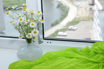 daisy flowers in a white vase on a white window-sill with bright green fabric and an open white window and street view with a river, road and cars