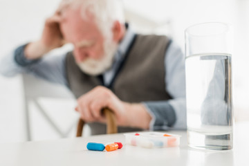 Selective focus of pills and water in glass on sad senior man background
