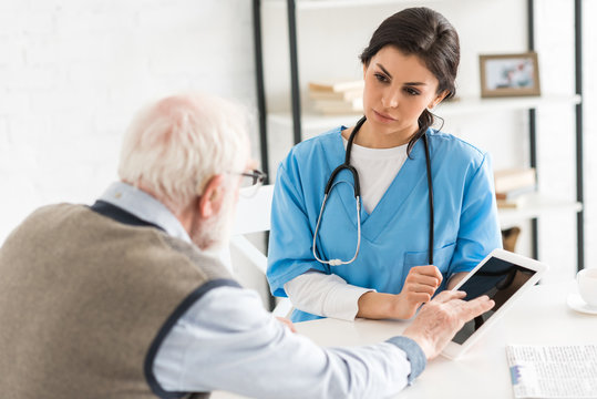 Retired Man Using Digital Tablet, And Sitting With Serious Doctor In Bright Room