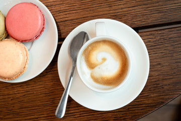 Espresso macchiato in white ceramic cup with saucer and spon next to a plate with macarons on brown wood surface from above.