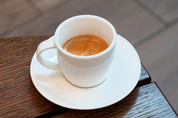 Italian espresso in a white ceramic cup with saucer on a brown wood table. Brurred background.