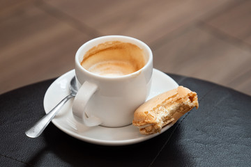 Half full cup of espresso macchiato with metal spoon and half eaten vanilla macaron on saucer. Black leather table.