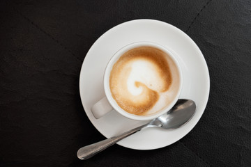 Espresso macchiato in white ceramic cup with saucer metal spoon isolated on black leather surface from above. Space for text.