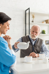 Cheerful nurse sitting on kitchen at home, while senior man pouring tea in cup