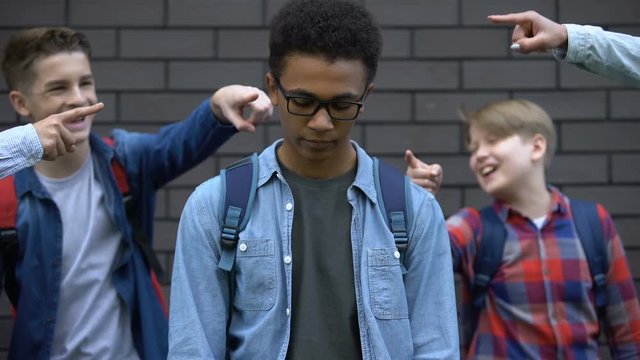 Group of cruel teenagers pointing fingers at black boy, humiliation and racism