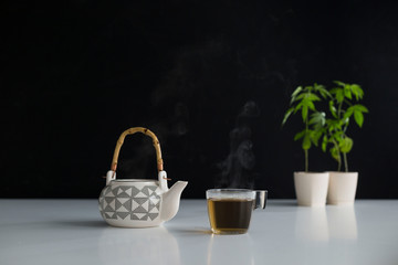 White table with Marijuana plant in pottery. Hot CBD tea in glass and teapot.