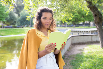 Positive young woman reading book in city park. Lady standing with blurred tree and river in...
