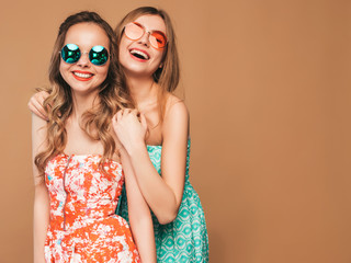 Two young beautiful smiling hipster girls in trendy summer colorful dresses. Sexy carefree women posing on golden background. Positive models going crazy in sunglasses