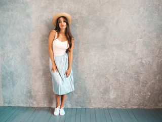 Portrait of Young beautiful woman looking at camera. Trendy girl in casual summer dress and hat. Funny and positive female posing near gray wall in studio