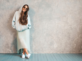 Young beautiful smiling woman looking at camera. Trendy girl in casual summer hoodie and skirt clothes. Funny and positive female posing near gray wall in studio. In sunglasses.