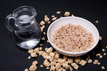 Bol con proteína de soja texturizada fina y gruesa rehidratada junto a una jarra con agua en fondo negro.