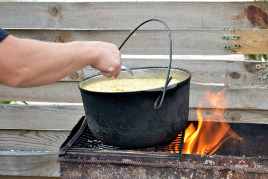 A man's hand mixes by spoon millet porridge in a pot on the grill with fire, on the wooden fence background