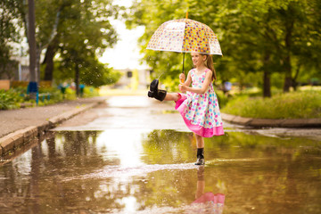 Happy funny kid girl with umbrella jumping on puddles in rubber boots and in polka dot dress and laughing