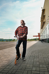 An elderly man with long hair and a beard rides a scooter through the city streets. Check shirt and jeans.