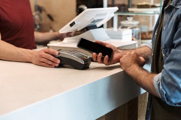 Cropped photo of young man paying debit card in cafe while waiter holding payment terminal