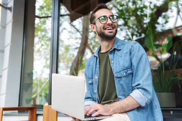 Photo of satisfied young man typing on laptop while working in city cafe outdoors