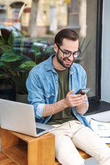 Photo of brunette young man holding smartphone while working on laptop in city cafe outdoors