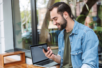 Photo of smiling young man holding smartphone while working on laptop in city cafe outdoors