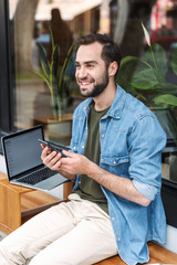 Photo of optimistic young man holding smartphone while working on laptop in city cafe outdoors