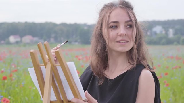 Portrait of cute smilling girl painting on the easel tanding on the poppy field. Young woman artist outdoors. Connection with nature. Love nature concept.