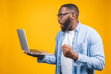 Young surprised african man standing and using laptop computer isolated over yellow background.