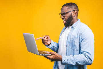 Young surprised african man standing and using laptop computer isolated over yellow background.