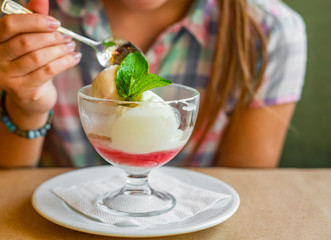 young girl's hand holding a spoon and  eating ice cream in glass at a table in cafe