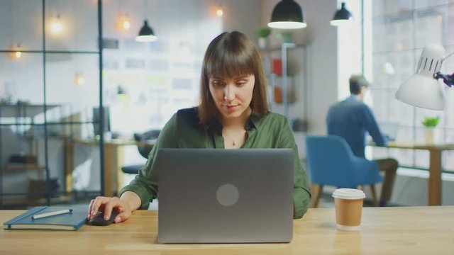 Young Beautiful Brunette Works on a Laptop Computer in Cool Creative Agency in a Loft Office. She has a Take-away Coffee and a Notebook on Her Table. Her Colleague Works in the Background. 