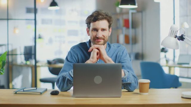 Young Handsome Man Works on a Laptop Computer in Cool Creative Agency in Loft Office. He has a Take-away Coffee and a Notebook on the Table. He Wears a Jeans Shirt. He Looks at the Camera and Smiles.