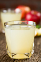 Refreshing apple juice in glass with apples in the back, photographed on rustic wood (Selective Focus, Focus on the front of the glass rim)