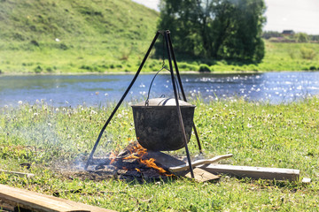 Cooking in a big tub on a halt on a travel on the river bank to sunny summer day. A fire under a cauldron with food. There is a smoke.