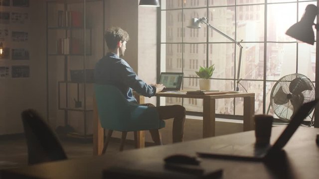 Handsome Creative Developer Works on a Laptop in His Studio. Designing Apps and Social Media Design Structure in His Sunny Loft Office. Modern Urban City View from the Window.