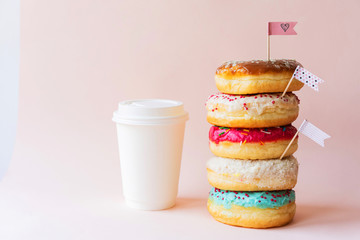 The colored glazed appetizing donuts are assembled in a tower and decorated with flag-shaped toppers, the paper cup is ideally white as a mock-up for text. Pink background with copy space. - obrazy, fototapety, plakaty